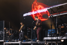 Na scenie występuje zespół muzyczny. Z przodu dwóch gitarzystów – jeden w garniturze, drugi w bordowym stroju. W tle perkusista za przezroczystą osłoną. Za nimi ekran z napisem „ARTYŚCI DLA TRANSP” i grafiką ręki z mikrofonem w czerwono-pomarańczowych barwach. Scena oświetlona reflektorami. Wokół instrumenty i sprzęt muzyczny.