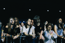 Grupa osób stoi za metalowym ogrodzeniem podczas koncertu. W tle ciemne niebo i jasne światła sceniczne. Większość patrzy w stronę sceny, niektórzy opierają ręce na barierce. Ubrania różnorodne – koszulki, bluzy. 