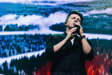 Mężczyzna w ciemnym garniturze śpiewa na scenie, trzymając mikrofon obiema rękami. Jego twarz jest zamazana. W tle duży ekran z zimowym lasem – drzewa pokryte śniegiem. Dominują kolory: niebieski, zielony i biały.