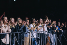 Grupa osób stoi za metalowym ogrodzeniem na koncercie. Większość ma uniesione ręce i klaszcze. Ubrania są różnorodne – od jasnych koszulek po ciemne kurtki. Tło jest ciemne, co wskazuje na noc lub słabe oświetlenie.