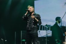 Mężczyzna w czarnej koszuli z białymi wzorami śpiewa na scenie. Trzyma mikrofon w prawej ręce. W tle duży ekran pokazuje jego powiększony wizerunek. Scena oświetlona zielonymi reflektorami.