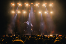 Na scenie trwa koncert. Wokalista stoi w jasnym świetle reflektorów, za nim ekran pokazuje jego powiększony obraz. W tle muzycy grają na instrumentach. Przed sceną tłum ludzi ogląda występ. Scenę oświetlają białe i żółte światła.