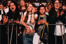 Grupa osób stoi za metalowym ogrodzeniem na koncercie. Twarze są zamazane. Jedna osoba trzyma telefon, inna ma plecak. W tle rozmyta publiczność. Ubrania w ciemnych kolorach.