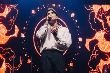 Mężczyzna w białej koszuli i ciemnych spodniach stoi na scenie z mikrofonem przy ustach. Wygląda, jakby śpiewał. Tło jest kolorowe, z pomarańczowo-czerwonymi wzorami przypominającymi chmury i gwiazdy. W centrum tła widoczny jest okrągły wzór otoczony jasnymi punktami.