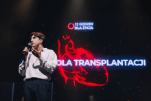 Mężczyzna w białej koszuli przemawia na scenie, trzyma mikrofon. W tle ciemny ekran z jasnym napisem „12 GODZIN DLA ŻYCIA” i „DLA TRANSPLANTACJI” na tle czerwonego rysunku serca.