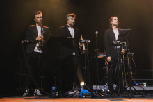 Na scenie stoi trzech muzyków w czarnych garniturach i białych koszulach. Grają na trąbce, saksofonie i puzonie. Przed nimi mikrofony na statywach i pulpity z nutami. Na podłodze butelki z wodą i sprzęt elektroniczny. Tło ciemne, oświetlone ciepłym, złotym światłem reflektorów.