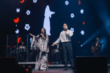 Na scenie śpiewa dwoje wokalistów. Kobieta po lewej ma długą, wzorzystą suknię kimono i trzyma mikrofon. Mężczyzna po prawej w białej koszuli i ciemnych spodniach, również z mikrofonem. W tle ekran z animacją: biała sylwetka kobiety, czerwone półksiężyce i białe kwiaty na czarnym tle. Widać też muzyków z instrumentami.