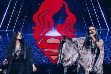 Na scenie stoją dwie osoby. Po lewej osoba w ciemnym stroju z naszyjnikiem trzyma mikrofon. Po prawej osoba w jasnym kimonie z kwiatowym wzorem gestykuluje ręką. W tle czerwony symbol Supermana i profil kobiety z długimi włosami na tle gwiaździstego nieba.