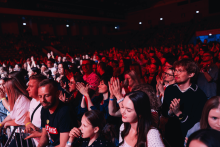 Tłum ludzi na koncercie. Wiele osób trzyma telefony, nagrywając lub robiąc zdjęcia. Scena oświetlona czerwonymi i niebieskimi światłami. Ludzie stoją blisko siebie, ubrani w jasne i ciemne stroje. W tle ciemna sala z punktowymi światłami.