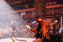 Na scenie koncertowej gitarzysta w czarnej kurtce i czapce stoi po prawej stronie. Po lewej wokalista trzyma mikrofon. Przed sceną tłum ludzi świeci latarkami telefonów. Scena oświetlona reflektorami. 
