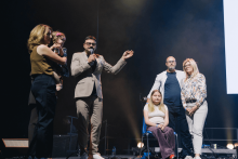 Na scenie znajduje się sześć osób. Jedna siedzi na krześle, pozostałe stoją wokół. Po lewej osoba trzyma dziecko. Mężczyzna w garniturze mówi do mikrofonu i gestykuluje. Tło ciemne, oświetlone jasnymi reflektorami. Po prawej częściowo widoczny duży ekran.