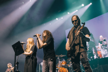 Na scenie występuje zespół muzyczny. W centrum dwie osoby śpiewają do mikrofonów – jedna z długimi blond włosami, druga z kręconymi ciemnymi. Po prawej gitarzysta w czarnej koszulce i szaliku w panterkę. W tle perkusista i duży ekran pokazujący obraz sceny. Jasne reflektory oświetlają scenę.