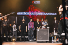Grupa osób stoi na scenie. W tle duży ekran z napisem „ARTYŚCI DLA TRANSPLANTACJI” oraz „12 GODZIN DLA ŻYCIA” i logo. Scena oświetlona ciepłym światłem. Uczestnicy ubrani są różnorodnie – od codziennych ubrań po stroje formalne. Po prawej stronie widoczny sprzęt nagłośnieniowy.