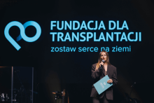 Kobieta trzyma mikrofon i niebieską teczkę z logo Fundacji dla Transplantacji. W tle duże logo fundacji i napis „Fundacja dla Transplantacji zostaw serce na ziemi” w jasnoniebieskim kolorze na ciemnym tle.