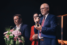 Na scenie stoją trzy osoby. Po prawej mężczyzna w granatowym garniturze i bordowym krawacie trzyma mikrofon. W środku osoba w czerwonym stroju, także z mikrofonem. Po lewej mężczyzna w ciemnym garniturze trzyma bukiet różowych i zielonych kwiatów. Tło ciemne, scena oświetlona reflektorami.