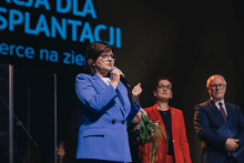 Kobieta w niebieskim garniturze przemawia na scenie, trzyma mikrofon. W tle dwie osoby – jedna w czerwonym stroju, druga w ciemnym garniturze. Za nimi napis „FUNDACJA DLA TRANSPLANTACJI Serce na ziemi” w kolorze niebieskim.