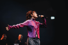 Osoba w fioletowej kurtce Adidas śpiewa na scenie, trzyma mikrofon w prawej ręce i ma rozłożone ramiona. W tle dwóch muzyków, jeden gra na klawiszach. Scena ciemna, oświetlona punktowym światłem.