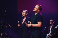 Na scenie śpiewa dwóch wokalistów. Kobieta po lewej w ciemnym ubraniu trzyma mikrofon w prawej ręce. Mężczyzna po prawej w czarnej koszulce również trzyma mikrofon. W tle gitarzysta z białą gitarą i statyw z nutami. Scena oświetlona fioletowym światłem.