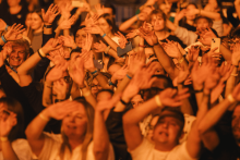 Tłum ludzi na koncercie z uniesionymi rękami. Niektórzy trzymają telefony. Uczestnicy mają na nadgarstkach opaski identyfikacyjne i różne ubrania. Twarze są zamazane. Tło rozmyte, oświetlenie w ciepłym, pomarańczowym odcieniu.