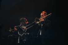 Dwaj muzycy grają na instrumentach dętych blaszanych. Po lewej trębacz, po prawej puzonista. Obaj ubrani w ciemne stroje. Stoją na scenie z ciemnym tłem. Twarz puzonisty zasłonięta kwadratem.