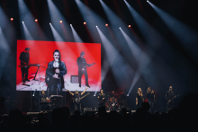 Na scenie występuje zespół muzyczny. Muzycy grają na gitarach, klawiszach i perkusji. Wokalista stoi z przodu. W tle duży ekran z czerwonym tłem pokazuje obraz członków zespołu. Scena oświetlona jasnymi reflektorami skierowanymi w dół.