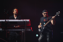 Na scenie dwóch muzyków. Jeden gra na białym keyboardzie z czerwonymi elementami, drugi na czarnej gitarze basowej. Tło ciemne, scena oświetlona punktowym światłem.