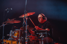 Mężczyzna gra na drewnianej perkusji z metalowymi elementami. Ma na sobie czarną koszulkę i słuchawki. Znajduje się na scenie. W tle widoczne jest ciemne oświetlenie sceniczne.