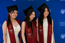 Trzy kobiety w białych sukniach i czarnych biretach stoją przed niebieską ścianą z napisem „Warszawski Uniwersytet Medyczny”. Mają bordowe szarfy z napisami „CLASS OF 2025” i „GRADUATE”.