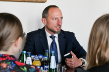 Mężczyzna w garniturze siedzi przy stole z butelkami napojów. Po bokach widoczne są sylwetki dwóch osób, uchwycone od tyłu.