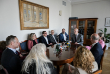 Kilka osób siedzi przy owalnym stole w eleganckiej sali spotkań z napojami na stole i książkami w tle.