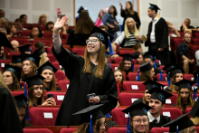 Grupa absolwentów w czarnych togach i biretach siedzi w czerwonych fotelach na sali. W centrum stoi osoba z telefonem w jednej ręce, machająca drugą. W tle widoczni są inni uczestnicy ceremonii, część z nich stoi.