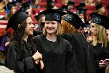 Grupa absolwentek w czarnych togach i biretach z fioletowymi chwostami stoi w sali. Na biretach widoczne jest niebieskie logo. W tle osoby siedzą na czerwonych krzesłach.