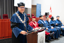 Uroczystość czepkowania absolwentów pielęgniarstwa i położnictwa. Na pierwszym planie stoi osoba w todze i birecie, przemawiająca przy mównicy z mikrofonem. W tle siedzi kilka innych osób również ubranych w togi.