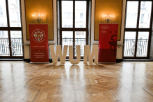 Elegancka sala z drewnianą podłogą i dużymi oknami. Na środku stoją duże, białe litery „WUM”. Po bokach dwa czerwone roll-upy z napisem „Warszawski Uniwersytet Medyczny Wydział Lekarski”. Na jednym herb uczelni, na drugim grafika kielicha.