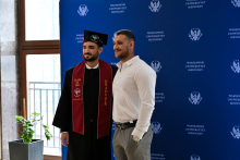 Dwóch mężczyzn pozuje na tle niebieskiej ścianki z logo Warszawskiego Uniwersytetu Medycznego. Jeden ma togę, czarny biret i bordową szarfę z napisem „Class of 2023” i „Graduate”. Drugi ma białą koszulę i szare spodnie. W tle okno i zielona roślina w doniczce.