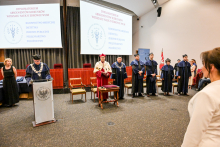 Grupa osób - rektor, dziekan i prodziekani Wydziału Nauk o Zdrowiu, w togach stoi na scenie podczas dyplomatorium wydziału. Za nimi dwa ekrany projekcyjne z napisem „Dyplomatorium Absolwentów Kierunków Wydziału Nauk o Zdrowiu WUM”. Po lewej mównica z herbem uczelni. Na pierwszym planie osoba patrzy na scenę.