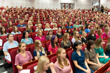 Uroczystość czepkowania absolwentów pielęgniarstwa i położnictwa. Grupa młodych kobiet i mężczyzn siedzących w audytorium, ubranych w kolorowe uniformy medyczne: różowe, niebieskie, zielone i fioletowe.