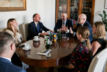 Siedem osób siedzi przy owalnym stole w sali konferencyjnej. Jedna z tych osób widoczna jest od tyłu. Na stole leżą dokumenty i stoją butelki z wodą. W tle znajdują się regały z książkami.