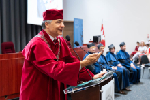 Uroczystość czepkowania absolwentów pielęgniarstwa i położnictwa. Na pierwszym planie stoi mężczyzna w czerwonej todze i birecie, mówi do mikrofonu. Mężczyzna ten nosi łańcuch rektorski. W tle siedzi kilka osób w niebieskich togach.