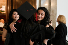 Dwie osoby obejmują się podczas ceremonii ukończenia szkoły. Jedna z nich ma czarną togę i czworokątną czapkę z czerwonym chwostem. W tle widoczne są inne osoby oraz wnętrze budynku z drewnianymi drzwiami i marmurowymi kolumnami.