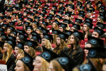 Duża grupa absolwentów w czarnych togach i biretach z niebieskim logo uczelni siedzi w czerwonych fotelach na sali audytoryjnej. Twarze osób są zamazane.
