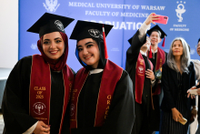 Grupa absolwentów Wydziału Lekarskiego Warszawskiego Uniwersytetu Medycznego. Na pierwszym planie dwie osoby w togach, biretach i bordowych szarfach z napisem „CLASS OF 2025” i „GRADUATE”. W tle niebieski baner z napisem „MEDICAL UNIVERSITY OF WARSAW FACULTY OF MEDICINE GRADUATION”. Inni robią selfie.