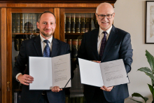 Dwaj mężczyźni w garniturach stoją przed regałem z książkami, trzymając otwarte teczki z dokumentami.