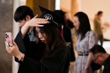 Młoda kobieta ubrana w czarną togę poprawia sobie biret patrząc w komórkę. W tle widać inne osoby, uczestników wydarzenia.