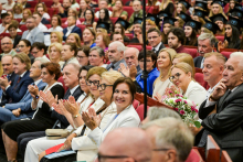 Duża grupa osób siedzi w audytorium. W pierwszym rzędzie osoby w eleganckich strojach trzymają bukiety kwiatów. W tle widać absolwentów w czarnych togach i biretach. Większość osób klaszcze.