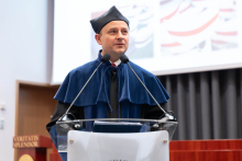 Uroczystość czepkowania absolwentów pielęgniarstwa i położnictwa. Mężczyzna stoi przy mównicy, przemawia do publiczności. ubrany jest w niebieską togę i biret.