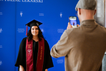 Absolwentka w czarnej todze i birecie pozuje do zdjęcia na tle niebieskiej ścianki z napisem „Wydział Lekarski” i „Warszawski Uniwersytet Medyczny”. Ma bordową szarfę z napisami „Class of 2025” i „Graduate”. Przed nią stoi fotograf z telefonem.