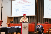 Rektor w czerwonej todze akademickiej z białym kołnierzem przemawia przy mównicy. Obok siedzi druga osoba w stroju akademickim. W tle ekran z napisem „Dyplomatorium Absolwentów Kierunków Wydziału Nauk o Zdrowiu WUM” i listą kierunków.