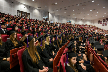 Duża sala pełna absolwentów w czarnych togach i biretach. Siedzą na czerwonych krzesłach, zwróceni w stronę sceny. W tle białe panele i obrazy. Na środku sali stoją osoby, prawdopodobnie goście.
