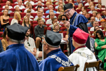 Na pierwszym planie cztery osoby w togach i biretach, jedna z czerwonym biretem. Na środku mężczyzna, dziekan, w niebieskiej todze przemawia do mikrofonu. W tle publiczność na czerwonych krzesłach.